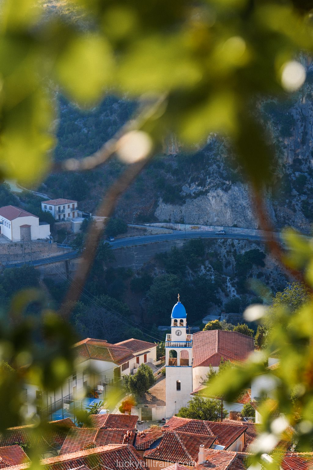 A church bell tower with a blue dome in the village of Dhërmι, seen through tree branches