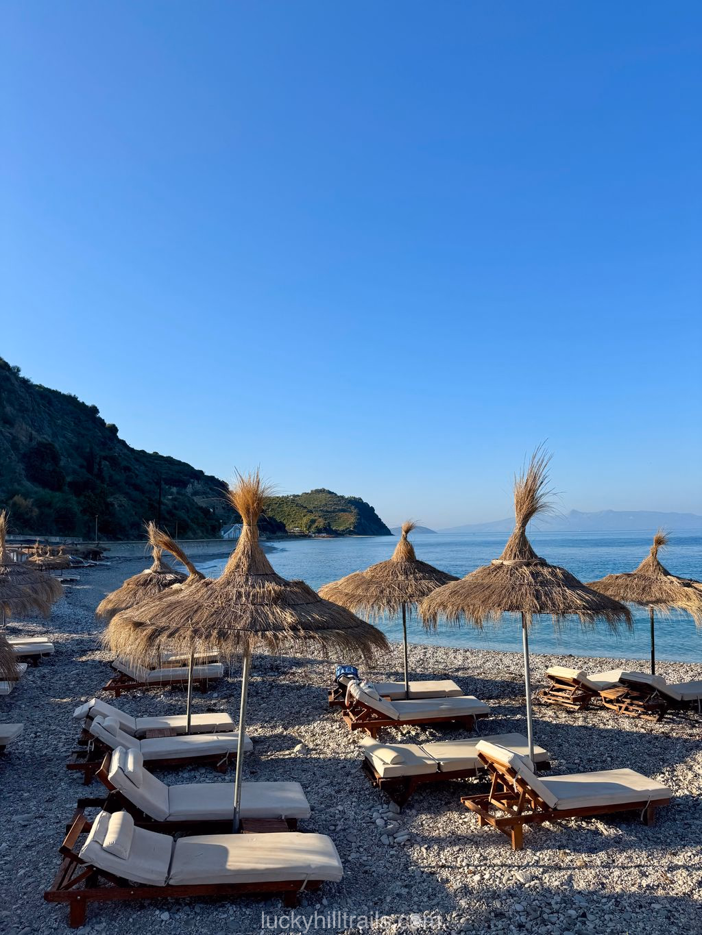 Plazhi i Bunec beach in Albania: sun loungers and straw parasols on a pebble shore, turquoise sea and green cliffs on the horizon