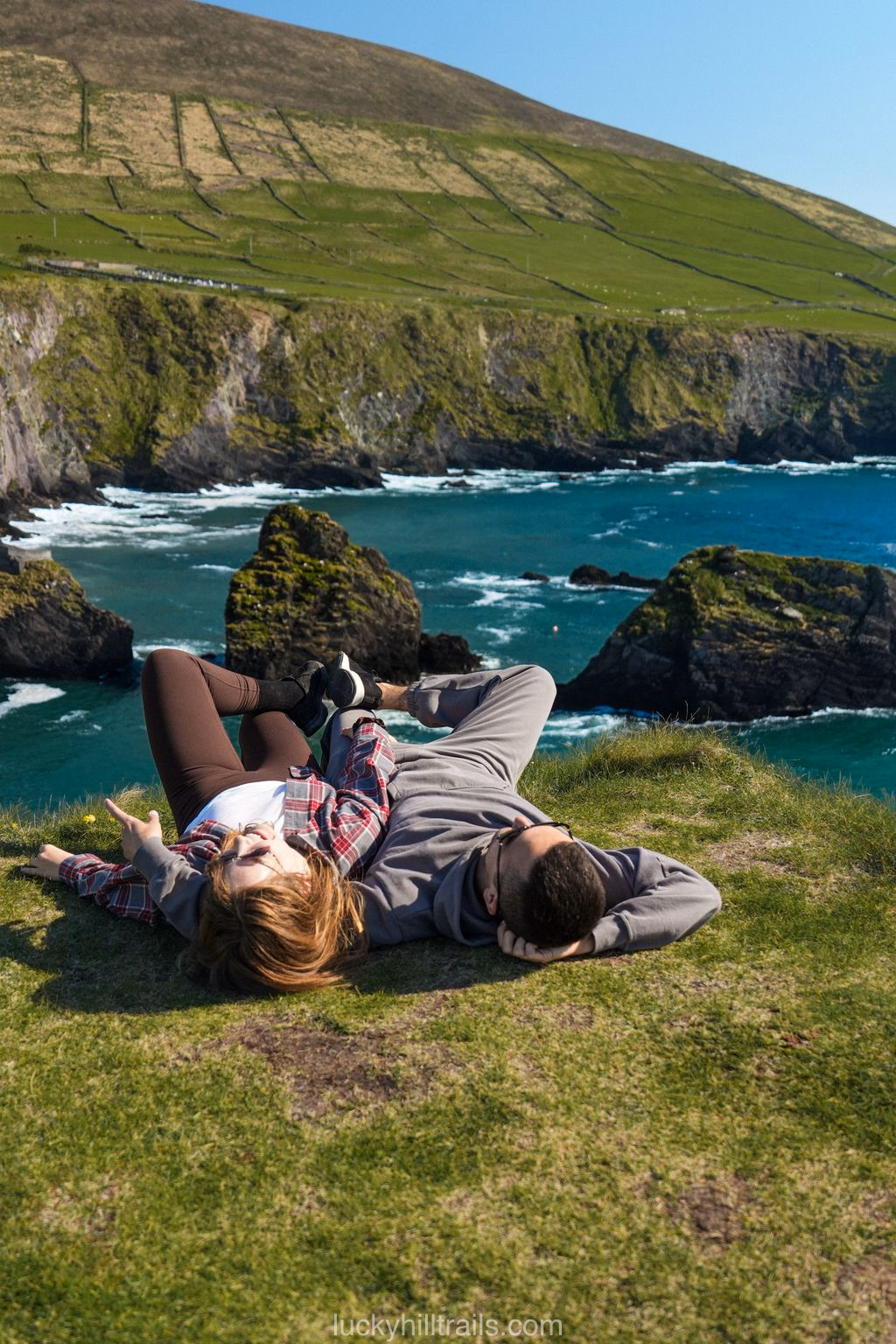 A couple rests on a green clifftop above the turquoise Atlantic with rocks below, Dingle Peninsula, Ireland