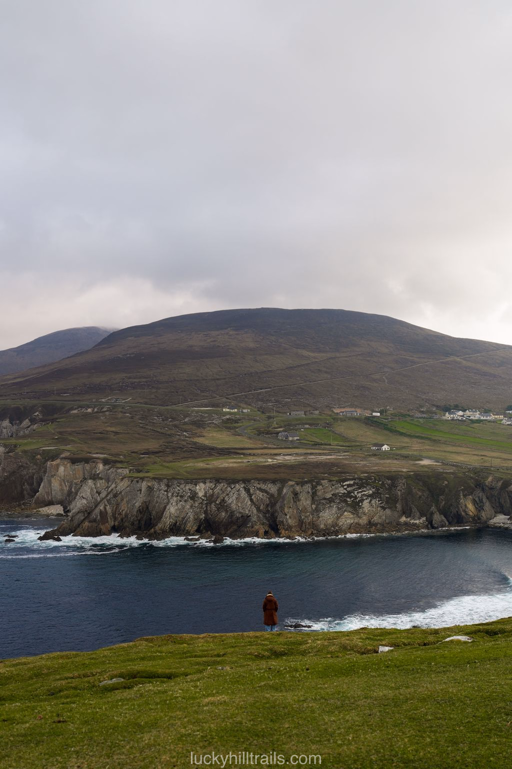A visitor stands at the edge of a green slope above the white Ashleam cliffs and the dark Atlantic, Achill Island, Ireland