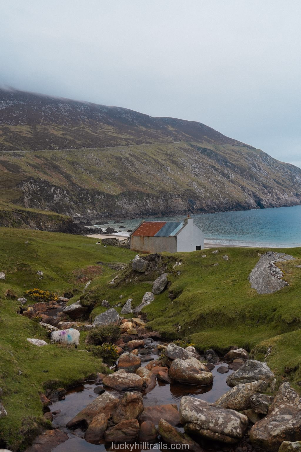 A stone cottage beside a stream with the turquoise Keem Beach bay and green mountains behind, Achill Island, Ireland