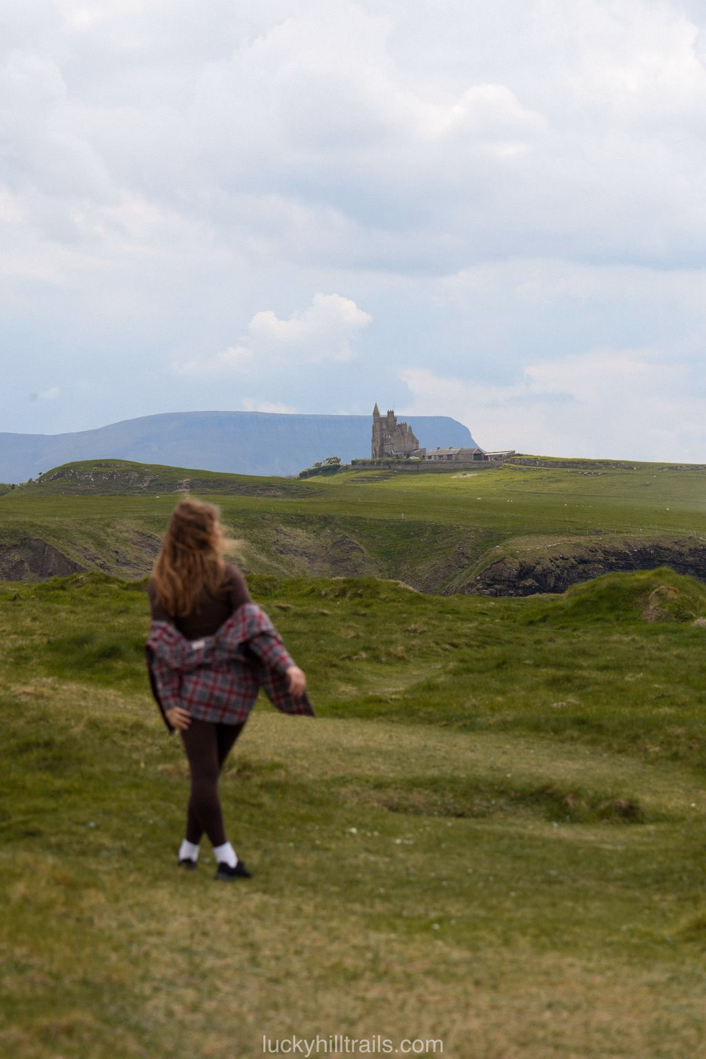A girl walks across green fields with Classiebawn Castle visible on the horizon, Mullaghmore, Sligo, Ireland