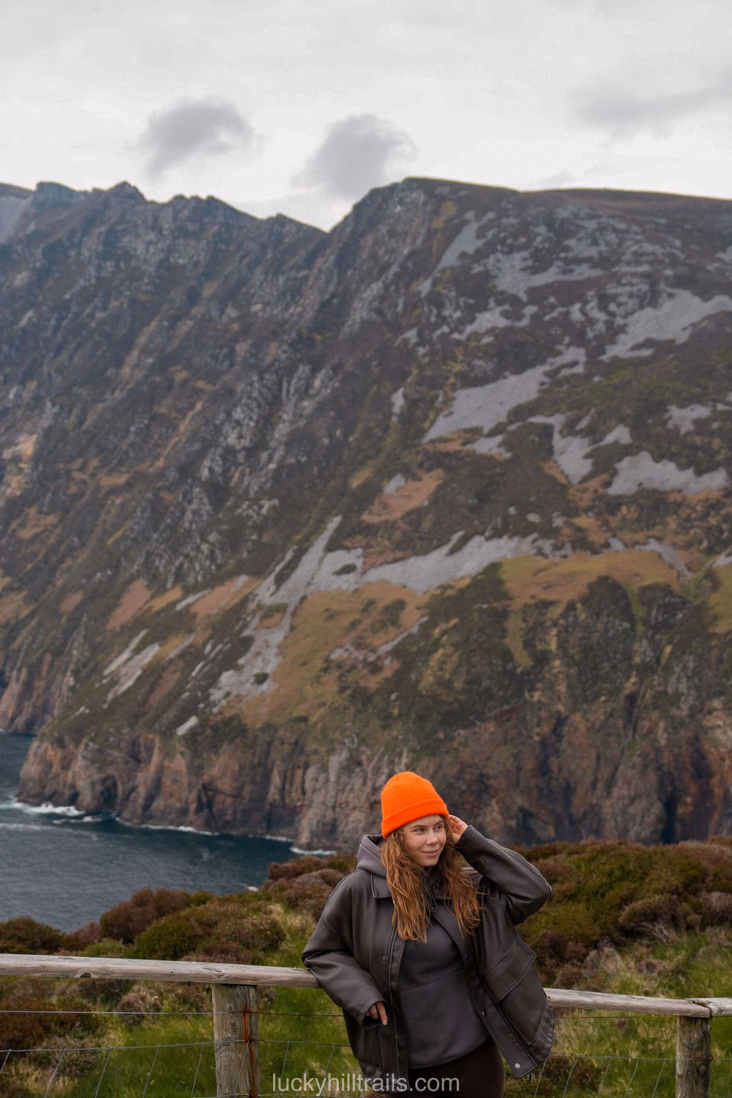 A girl in an orange beanie against the sheer drops of Slieve League above the dark ocean and heather in the foreground, Donegal, Ireland