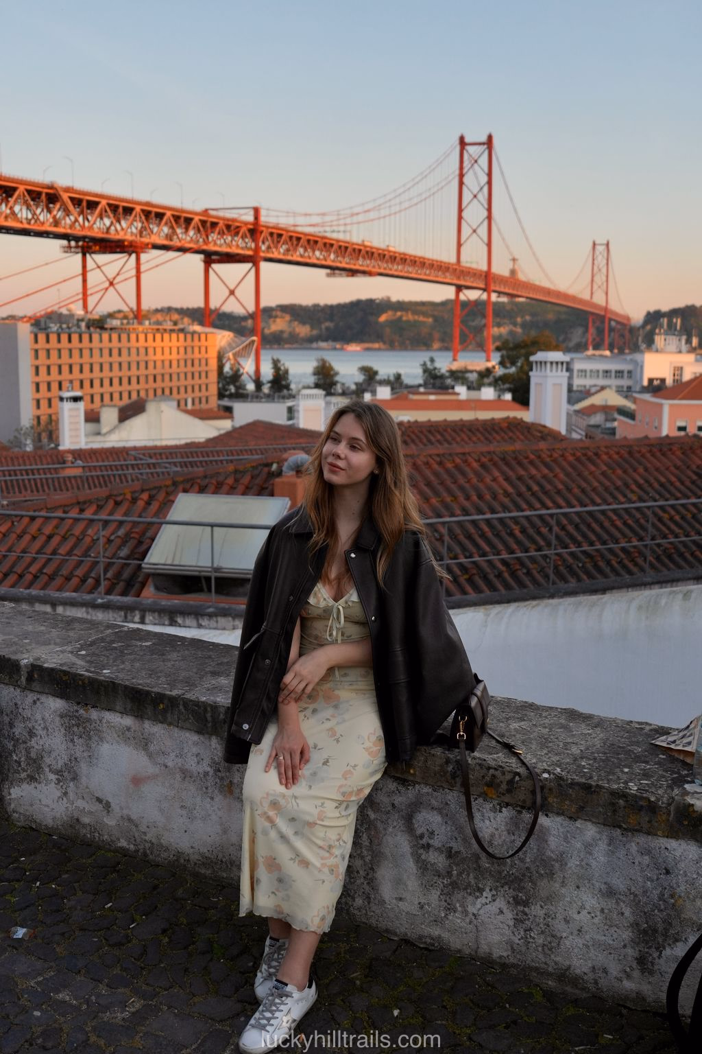 Girl in a floral dress and leather jacket at Miradouro de Santo Amaro – the red Ponte 25 de Abril bridge over the Tagus river in evening light behind her, Lisbon, Portugal