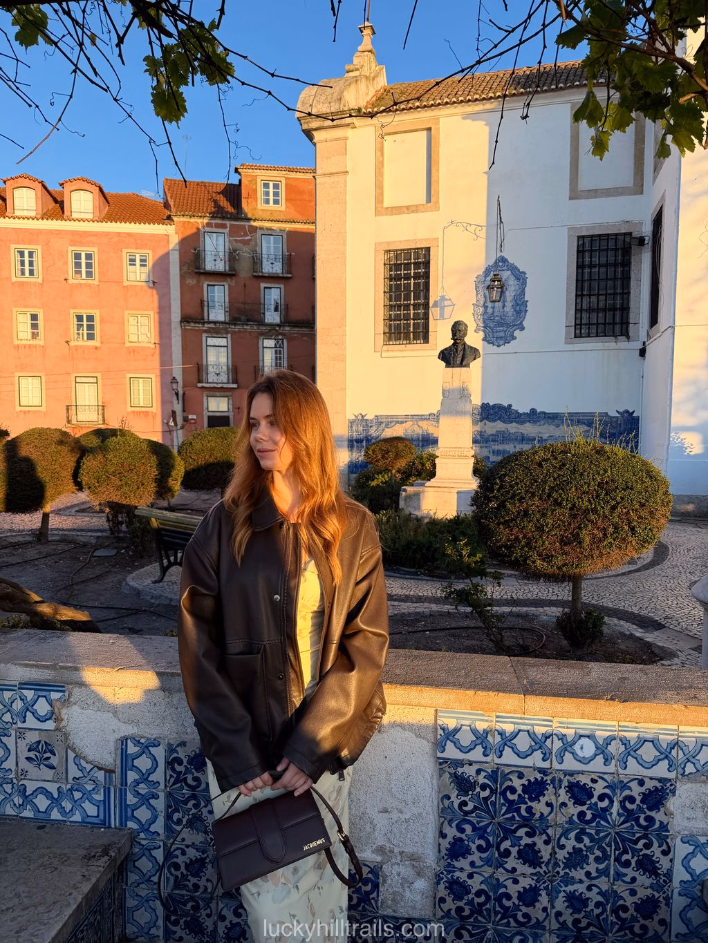 Girl in a brown leather jacket at Largo das Portas do Sol in golden evening light, white azulejo-tiled building and manicured garden in the background, Lisbon, Portugal