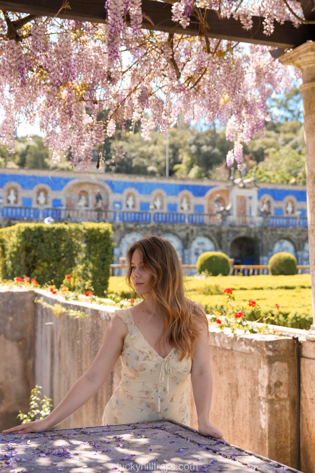 Girl in a floral dress standing under hanging wisteria clusters in the gardens of Palace Fronteira, azulejo-decorated gallery in the background, Lisbon, Portugal