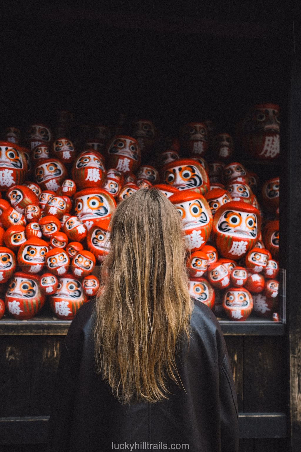 Girl standing with her back to a wall of red daruma dolls at Katsuo-ji Temple