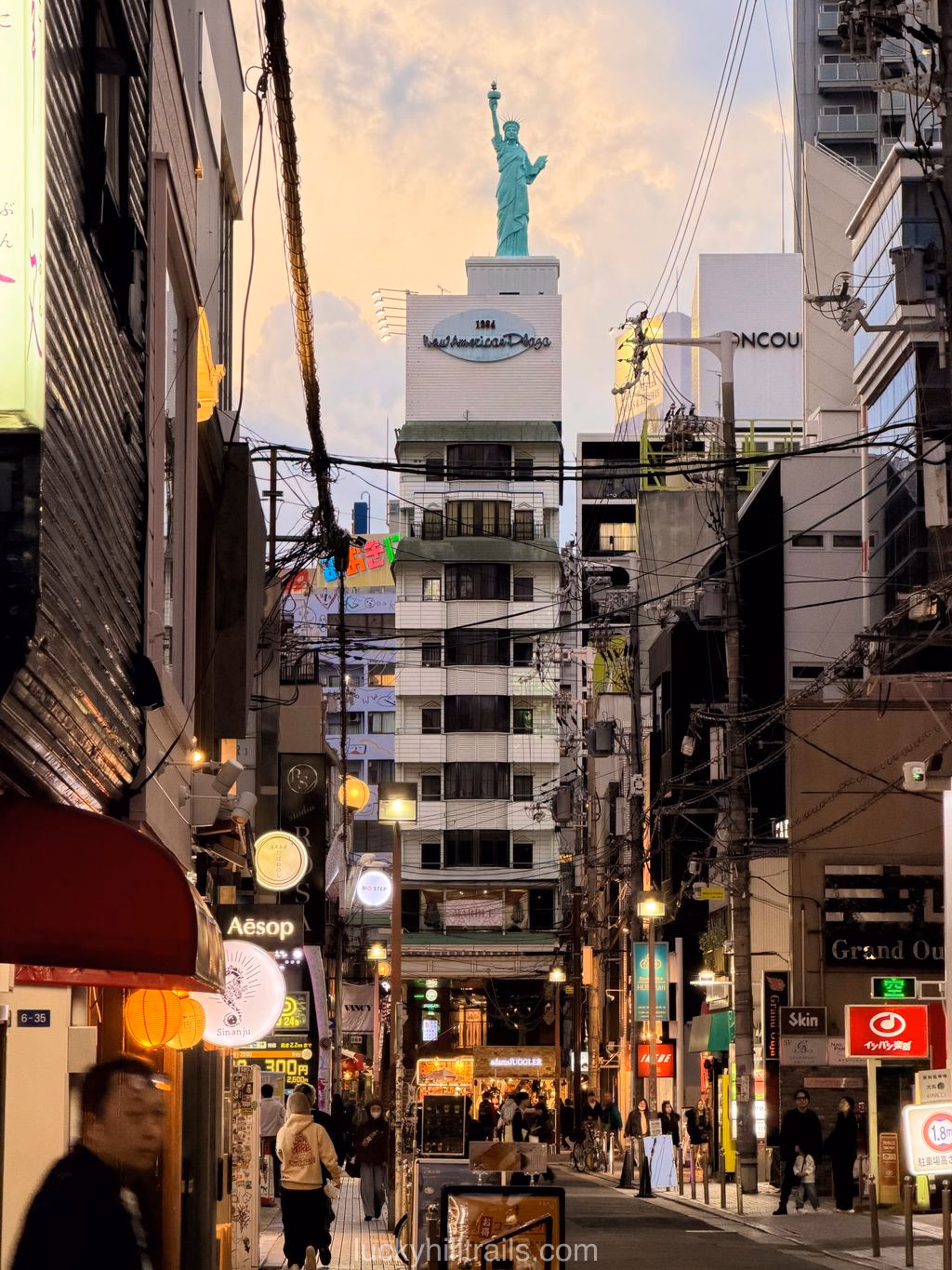 A narrow street in the American Village district of Osaka in the evening: shops, signs, and a Statue of Liberty on a rooftop in the distance