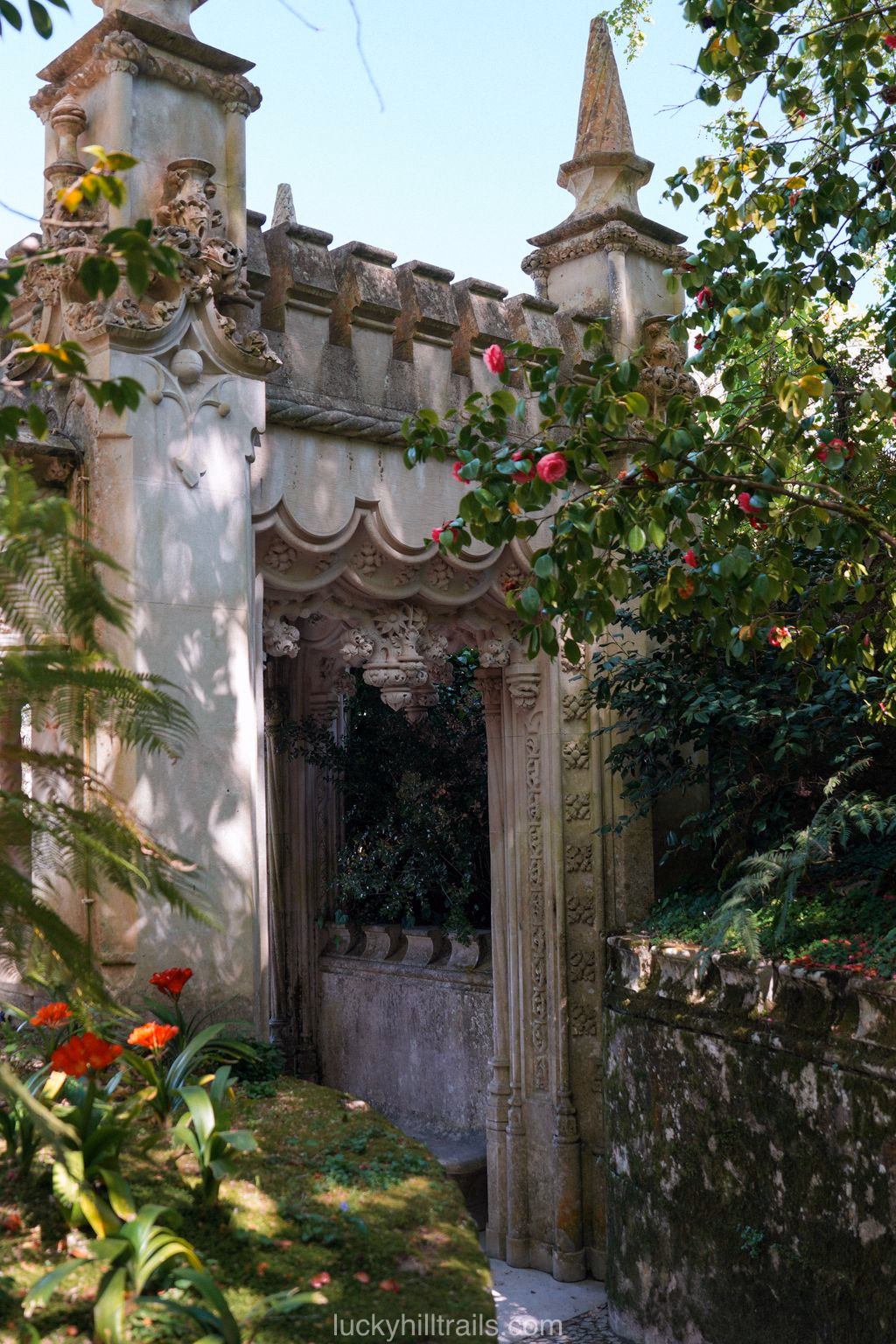 Gothic garden arch with towers and ornate stonework in the grounds of Quinta da Regaleira, surrounded by flowering roses and greenery, Sintra, Portugal